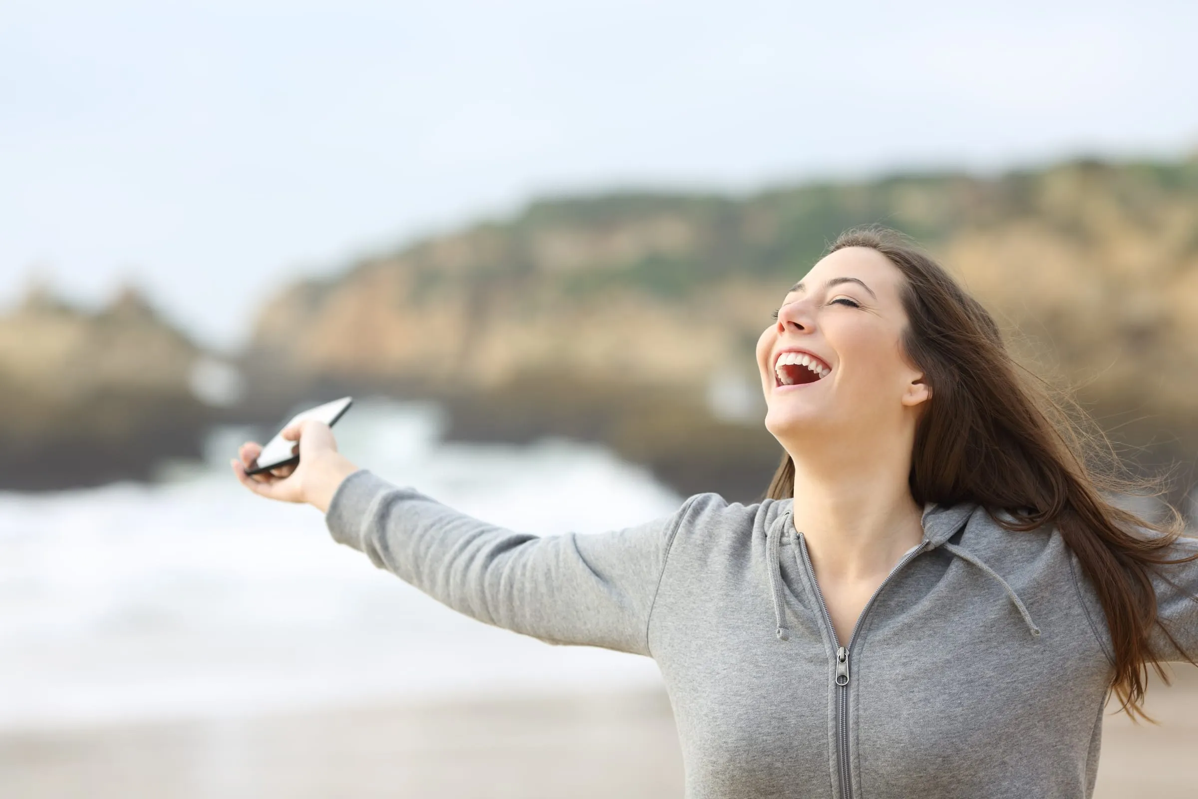 A woman laughing as she reads good news on her phone at the beach
